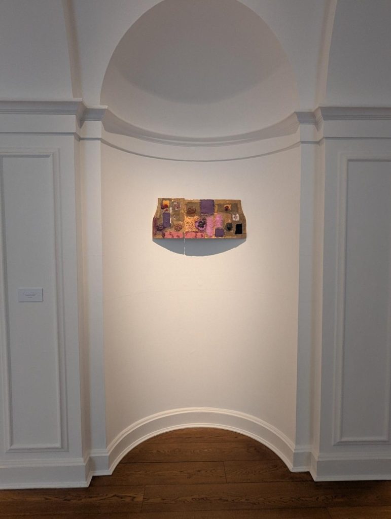 An artwork by Joan Snyder displayed on a wall in an elegant gallery setting, featuring textured and colorful elements arranged in a wavy rectangular shape.