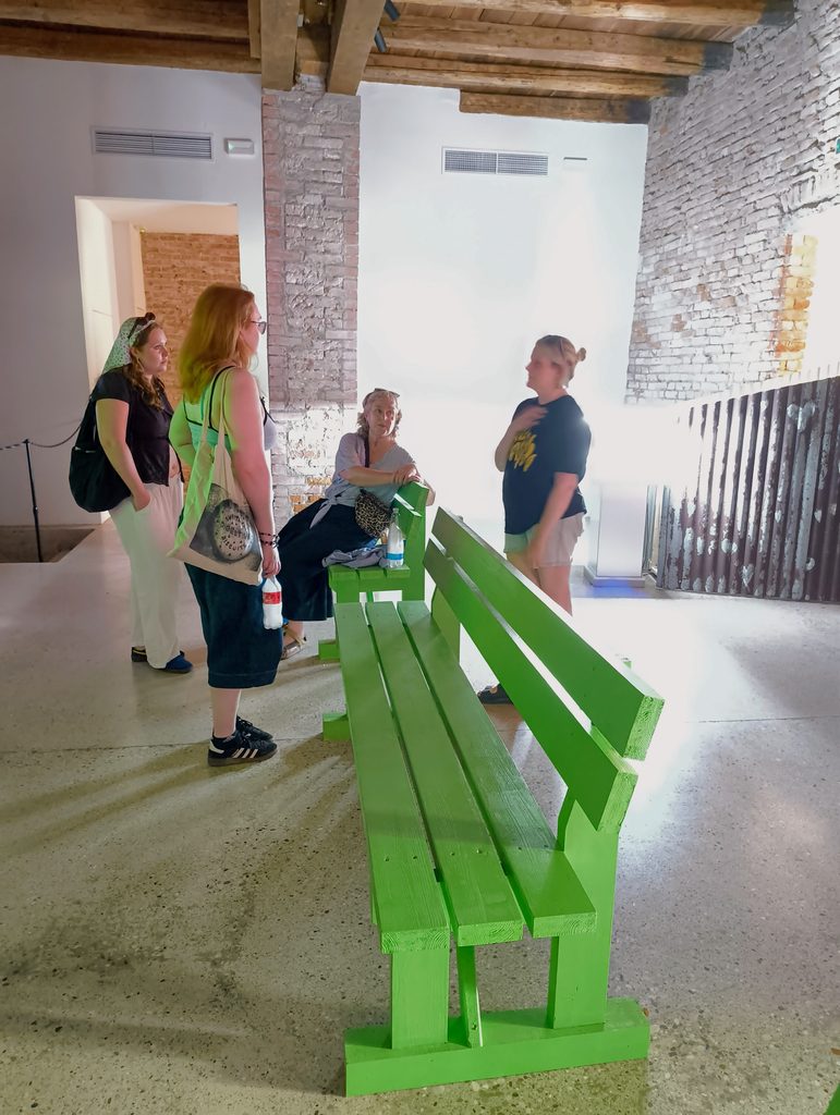 Visitors gather around a bright green bench installation inside a gallery space with exposed brick walls, timber ceiling beams and polished concrete floors.