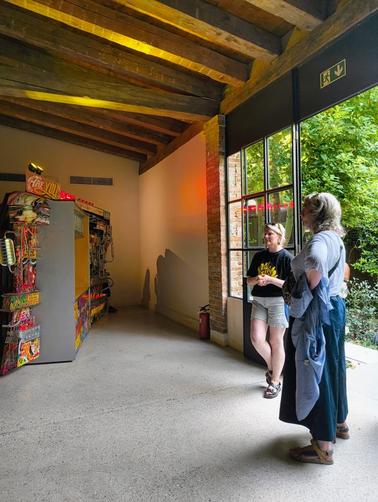 Visitor and guide observing a large-scale mixed-media installation in a gallery space, with illuminated sculptural elements, exposed timber ceilings and natural light entering through an open doorway to an outdoor garden.