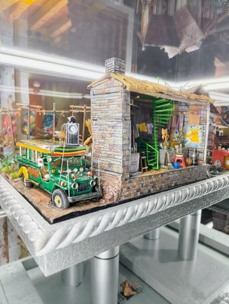 Detailed miniature sculpture showing a small brick-and-timber building beside a brightly decorated green jeepney. The model includes a green spiral staircase, hanging laundry, trophies, plants and various household objects displayed inside a glass case.
