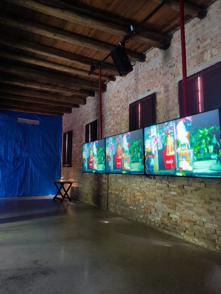Video installation displayed across four screens in a historic gallery space with exposed brick walls, timber ceiling beams and a blue fabric backdrop.