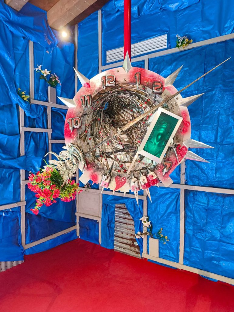 Mixed-media sculptural clock installation suspended within a blue-walled exhibition space, featuring decorative spikes, flowers and a red floor.
